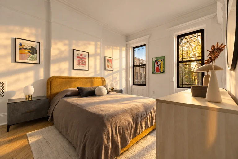 Golden-hour bedroom with a mustard headboard, gray bedding, framed art and two tall windows.