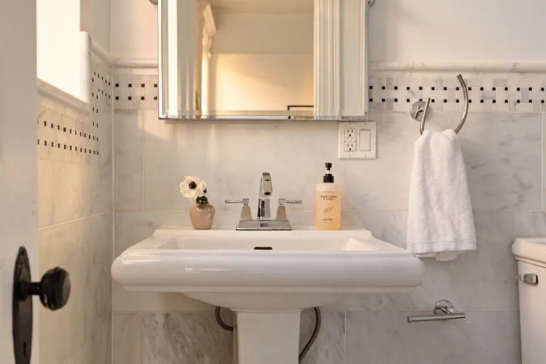 Pedestal sink, chrome faucet, white towel and marble tile in warm reflected afternoon light.